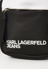 Svart nylon veske med glidelås, teksturert overflate og fremtredende hvit tekst som sier "KARL LAGERFELD JEANS."