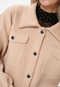 Beżowa, strukturalna kamizelka koszulowa z kołnierzykiem, czarnymi guzikami i dwiema kieszeniami na piersi. Nosić ją na czarną bluzkę z siateczki w kropki.