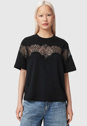 Femme aux longs cheveux blonds portant un haut noir à manches courtes avec un panneau transparent en dentelle florale sur la poitrine, assorti d'un jean bleu.