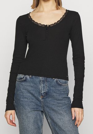 Vrouw draagt een zwarte top met lange mouwen, kanten rand en knopen, gecombineerd met een high-waisted blauwe spijkerbroek, staand tegen een eenvoudige achtergrond.