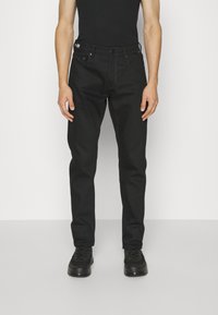 Jeans in denim nero con vestibilità dritta, dotati di tasche frontali, chiusura a bottone e dettagli di cucitura sottili. Indossati con una canotta nera e sneakers.