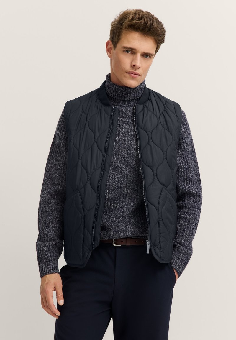 Gestepptes schwarzes Weste mit hohem Kragen und Reißverschluss. Über einem dunklen strukturierten Pullover getragen. Taschen sichtbar, mit glatter Stoffoberfläche.