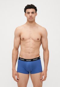 Blauwe boxershorts met een zwarte elastische tailleband waarop het merk "HUGO" in witte tekst staat. Gladde stof en een aansluitend ontwerp.