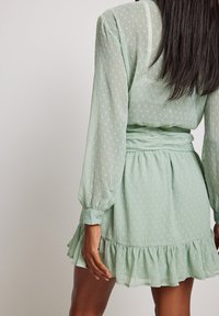 Abito verde menta con maniche lunghe a palloncino, vita arricciata e orlo con volant. Il tessuto presenta un motivo testurizzato sottile in tonalità più chiare.