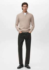 Maglione a costine beige con colletto a zip, abbinato a pantaloni dritti verdi scuri e scarpe nere. Design minimalista, superficie testurizzata.