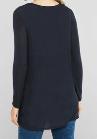 Žena v modré tunice s dlouhým rukávem a modrých džínsech, zobrazená zezadu na světlém pozadí.