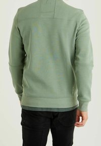 Uomo che indossa un felpa a maniche lunghe verde chiaro e pantaloni neri, visto da dietro su uno sfondo chiaro semplice.