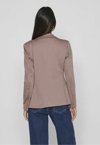 Blazer marron sur mesure à manches longues, texture lisse et coupe ajustée, associé à un jean bleu taille haute. Vue de dos.