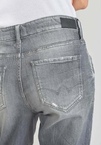 Harmaat farkut haalistuneella viimeistelyllä. Takana on taskut tunnistettavalla ompelukuviolla ja vyötäröllä musta brändilaatta.