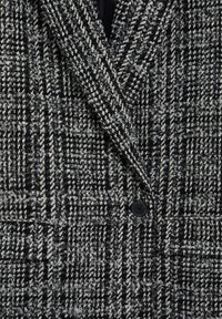 Blazer croisé en tweed noir et blanc, avec un col à revers cranté, un bouton noir, et un tissu texturé avec un léger éclat.