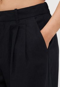 Czarne lniane spodnie z wysokim stanem, z lewą kieszenią, plisowanym przodem i gładką fakturą. Odpowiednie do strojów formalnych lub codziennych.