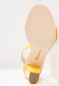 Sandale à talon haut jaune avec talon bloc, bout ouvert, bride à la cheville et semelle beige indiquant la marque "Tamaris" et la pointure 37.