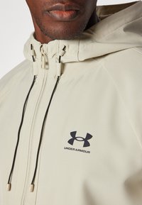 Giacca sportiva beige con zip frontale, cappuccio regolabile e logo Under Armour nero. Presenta cordini con fermagli in plastica per adattarsi.