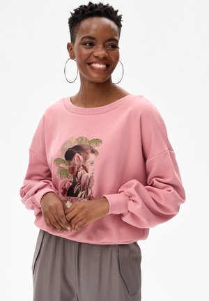 Lachende vrouw met kort krullend haar, grote ronde oorbellen, een roze sweatshirt met bloemmotief en grijze broek tegen een witte achtergrond.