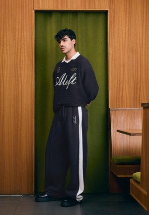 Młody mężczyzna z kręconymi włosami stoi na tle zielonej zasłony, ubrany w czarny sweter "Aloft", czarne spodnie z białym paskiem i czarne buty w pokoju z drewnianą boazerią.