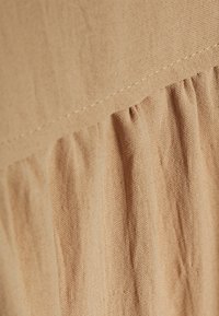 Bežová látka s texturovaným povrchem, která obsahuje volánkovou část. Šití je viditelné podél okraje a zdůrazňuje design oděvu.