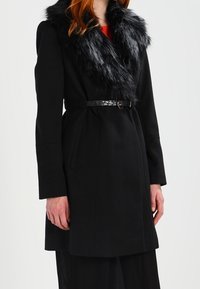 Svart kappa med krage av konstgjord päls, bälte i midjan och långa ärmar. Texturerat tyg med rak klippning som når mitt på låret.