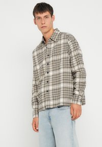 Ruudullinen plaid-paita beige, ruskea ja musta. Pitkähiha, napitettava etuosa, yksi rintatasku, tehty pehmeästä kankaasta. Yhdistetty vaaleansinisiin farkkuihin.