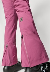 Roze skibroek met contrasterende ritsen, textuuraccenten en een logo-patch. Strakke pasvorm bij de enkels, gecombineerd met zwarte snowboots.