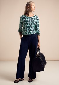 Grön och teal-mönstrad långärmad topp, marinblå vida byxor, svarta nät-slip-on skor, och en svart handväska. Släta texturer och avslappnad passform.