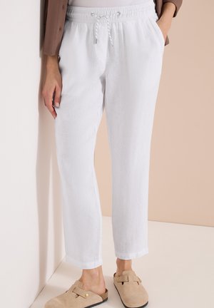 Personne portant un pantalon en lin blanc à cordon avec poches latérales et des chaussures beiges sans lacets, se tenant contre un mur beige clair.