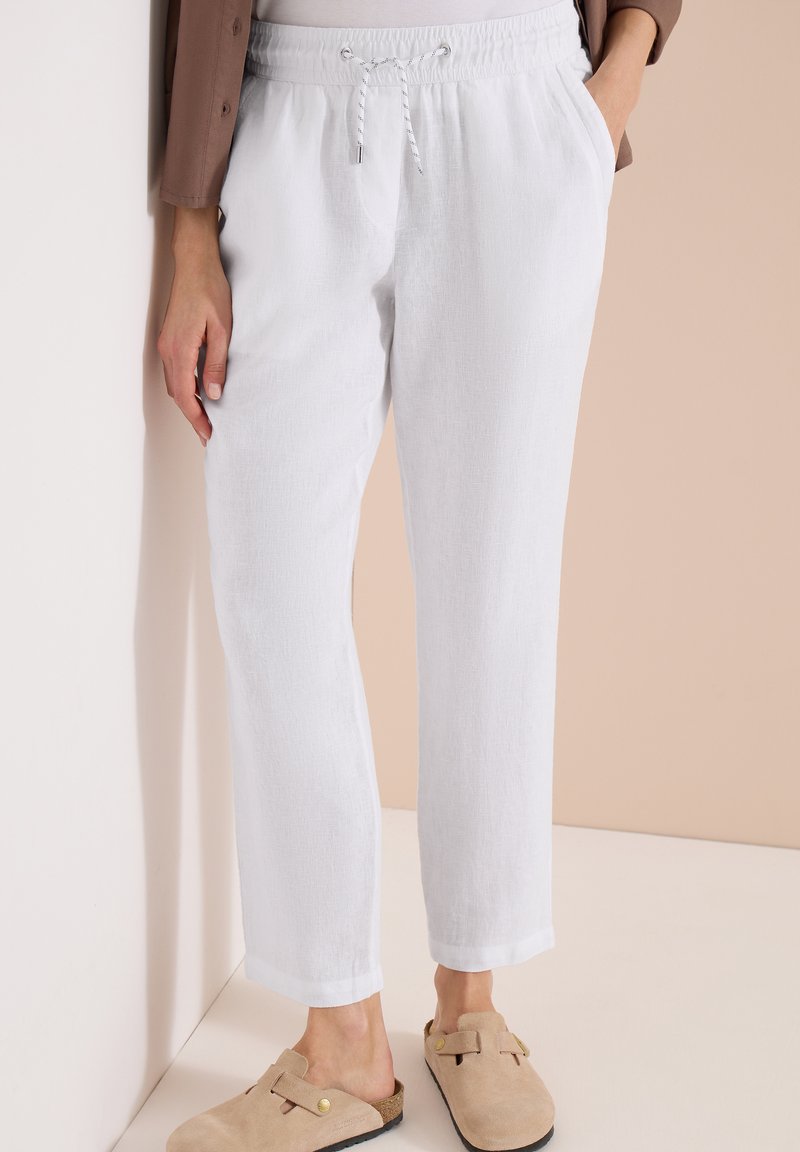 Personne portant un pantalon en lin blanc à cordon avec poches latérales et des chaussures beiges sans lacets, se tenant contre un mur beige clair.