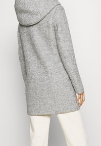 Grå ullrock med struktur, utrustad med huva och långa ärmar, som faller nedanför höfterna. Minimalistisk design och rena linjer.