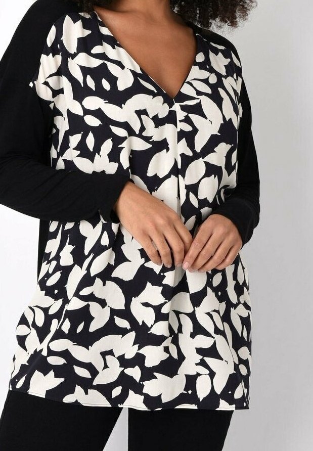 Woman wearing a black and white leaf-patterned long sleeve top with a V-neck, black sleeves, and black pants, hands gently clasped in front.