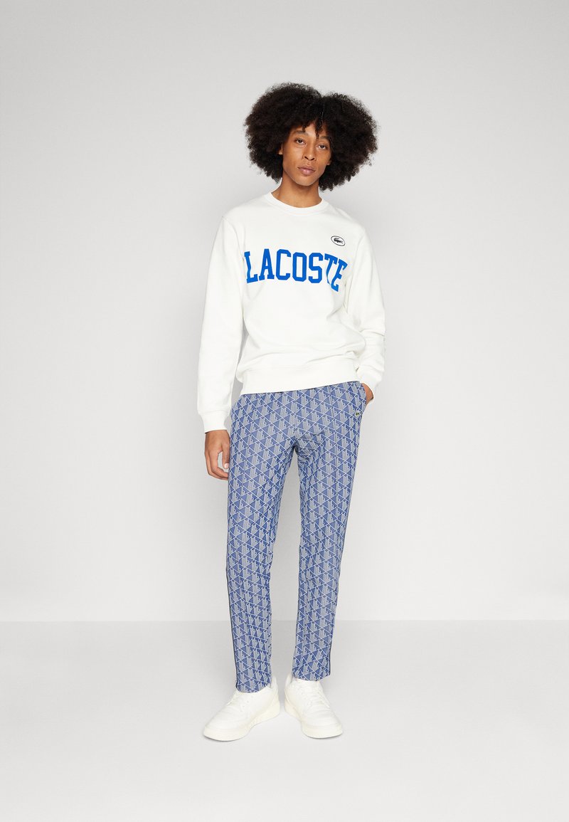 Felpa bianca con il testo "LACOSTE" blu, abbinata a pantaloni blu con motivo e sneakers bianche. Design semplice con polsini e vita a costine.