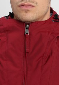 Rød zip-jakke med høj krave. Funktionerne inkluderer en sølvfarvet metallynlås og justerbare snore, lavet af et glat, let materiale.