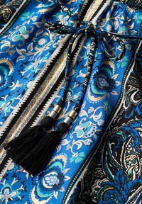 Tissu bleu avec motifs floraux et paisley, avec un cordon noir noué en nœud orné de perles métalliques et de pompons noirs.