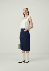 Hvit crop topp, mørkeblå denim midi-skjørt, hvite cowboystøvler og en liten grå håndveske. Modellen står mot en nøytral bakgrunn.