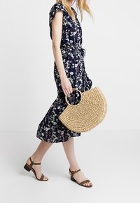 Blommigt mönstrad klänning i marinblått med korta ärmar och knytband i midjan; matchad med en vävd halvmåne-stråväska och bruna sandaler med remmar.