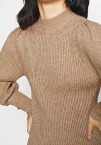 Ribbade, höghalssvetrar i beige, med puffade axlar och åtsittande ärmar, tillverkad av ett mjukt, texturerat stickat material.