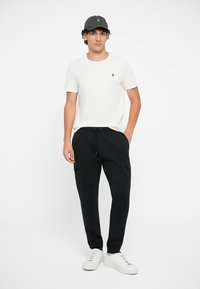 Camiseta de algodão branca, calças cargo pretas com bolsos e tênis brancos. Acessórios incluem um boné de beisebol cinza escuro. Roupa casual.