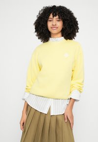 Żółta bluza z prążkowaną teksturą, z małym diamentowym logo. Do tego biała i szara pasiasta koszula oraz plisowana brązowa spódnica.