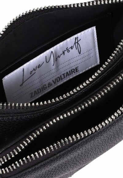 Sac en cuir noir avec des fermetures éclair argentées ouvertes dévoilant une étiquette blanche à l'intérieur portant les inscriptions "Love Yourself" et "Zadig & Voltaire".