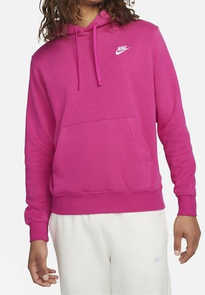 Osoba ubrana w jasnoróżową bluzę z kapturem Nike z przednią kieszenią i sznurkami, zestawioną z białymi spodniami dresowymi Nike.