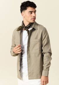 Beige skjorta med knappar, bröstficka, långa ärmar och mandarin-krage, tillsammans med en mörkgrön mönstrad scarf. Texturerat tyg.