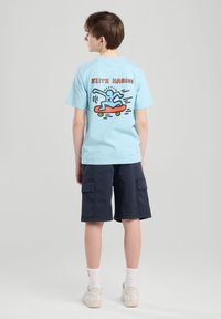 T-shirt in cotone azzurro chiaro con una grafica di uno skater sulla schiena, abbinato a pantaloni cargo scuri e scarpe da ginnastica bianche.