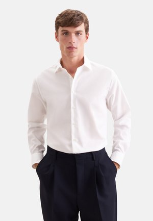 Jonge man die een eenvoudige witte blouse met lange mouwen en donkere geplooide broek draagt, met handen in de zakken tegen een effen achtergrond.