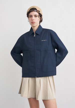 Młoda osoba ubrana w granatową kurtkę Carhartt na zamek, beżową plisowaną spódnicę i beżową czapkę z daszkiem założoną do tyłu na jednolitym tle.