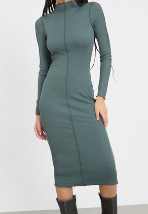 Vrouw draagt een aansluitende, lange mouwen, hoge hals, geribde midi-jurk in gedempt groen met een centrale naad en zwarte halfhoge laarzen.