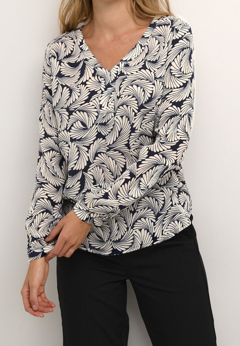 Femme portant un chemisier à manches longues bleu marine et blanc à motif de feuilles avec un col en V, ajustant sa manche, associé à un pantalon noir.