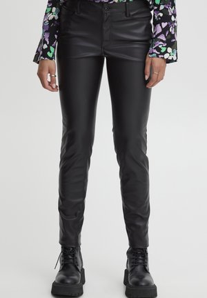 Pantaloni sintetici neri in pelle con vestibilità slim. Presentano una cerniera frontale, passanti per la cintura e una texture liscia e lucida. Abbinati a stivaletti militari neri.