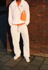 Vit jacka med orange detaljer över en orange skjorta, i kombination med vita byxor och sneakers. Liten orange handväska. Tegelvägg i bakgrunden.