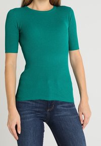 Teal ribbed short-sleeve top with a round neckline, made of stretchy material, paired with blue denim jeans.