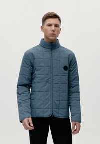 Chaqueta acolchada azul con un cuello alto, cremallera frontal y un bolsillo en el pecho con un acento negro redondo. El material parece ligero y texturizado.