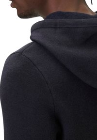 Felpa con cappuccio blu scuro con una texture morbida, caratterizzata da un cappuccio con coulisse e cuciture lisce lungo le spalle. È visibile una scollatura rotonda.