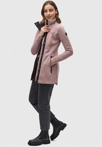 Manteau rose à manches longues avec col haut et accents noirs. Fermetures à glissière et à bouton-pression ; poches latérales. Assorti à un jean foncé et des bottes noires.